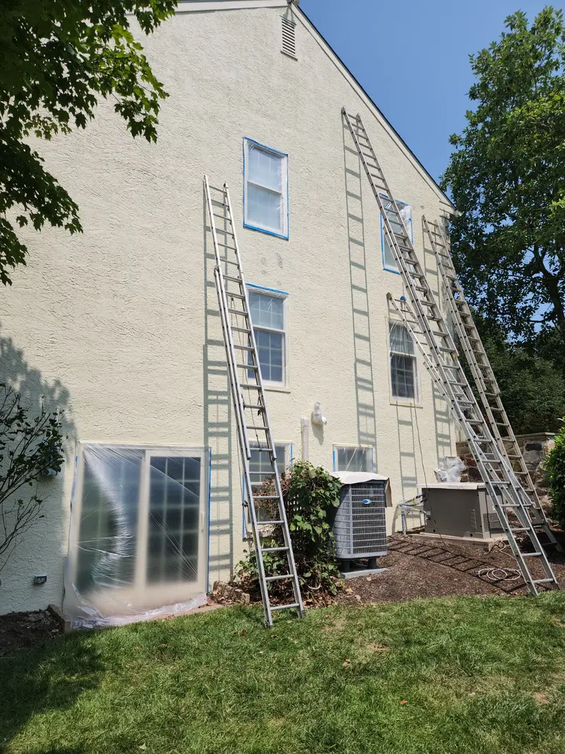 Exterior stucco painting in progress with ladders and window masking, Malvern, PA